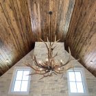Yellowstone Elk Antler Chandelier hanging from a vaulted wood ceiling above a stone chimney between two tall windows