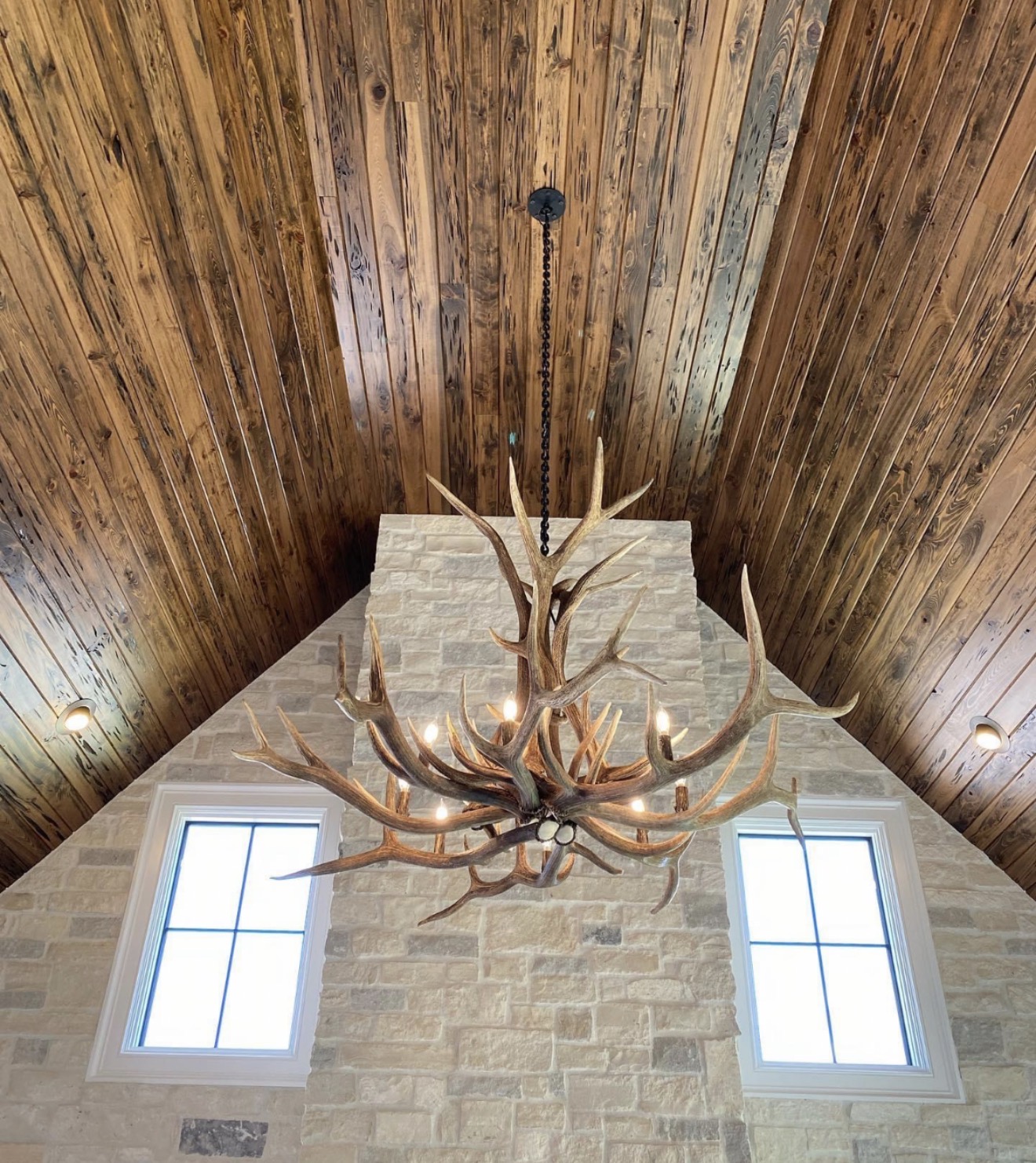 Yellowstone Elk Antler Chandelier hanging from a vaulted wood ceiling above a stone chimney between two tall windows