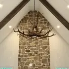 Wilderness Elk Antler Chandelier in a natural finish, hanging from a vaulted white ceiling with dark wood beams in front of a tall stone chimney