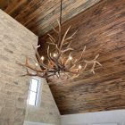 Yellowstone Elk Antler Chandelier hanging beneath a wood plank ceiling beside a stone chimney and window