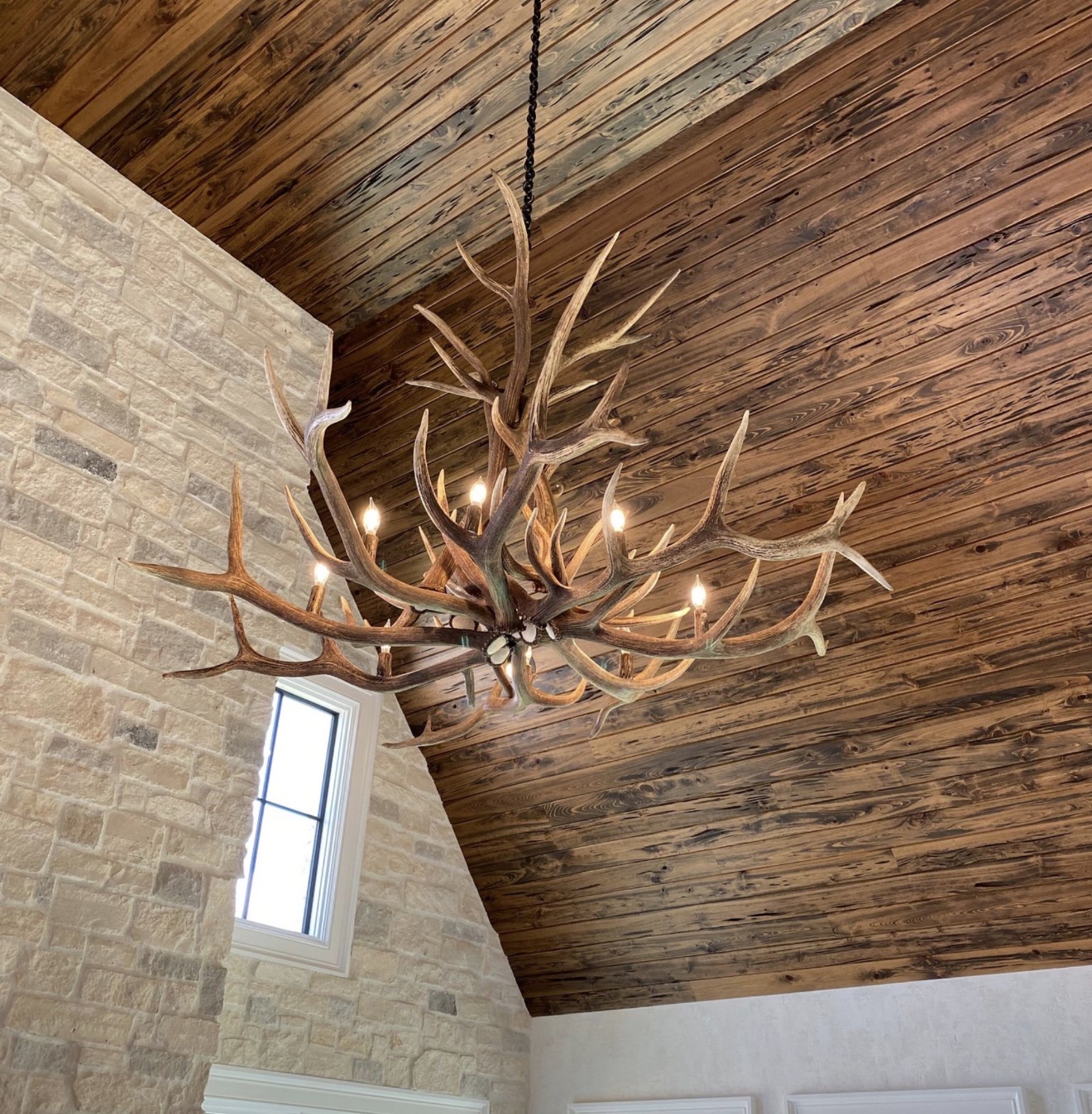 Yellowstone Elk Antler Chandelier hanging beneath a wood plank ceiling beside a stone chimney and window