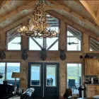 XL 6 tier whitetail antler chandelier installed over a vaulted great room with wood beams, mounted deer heads, and a lake view