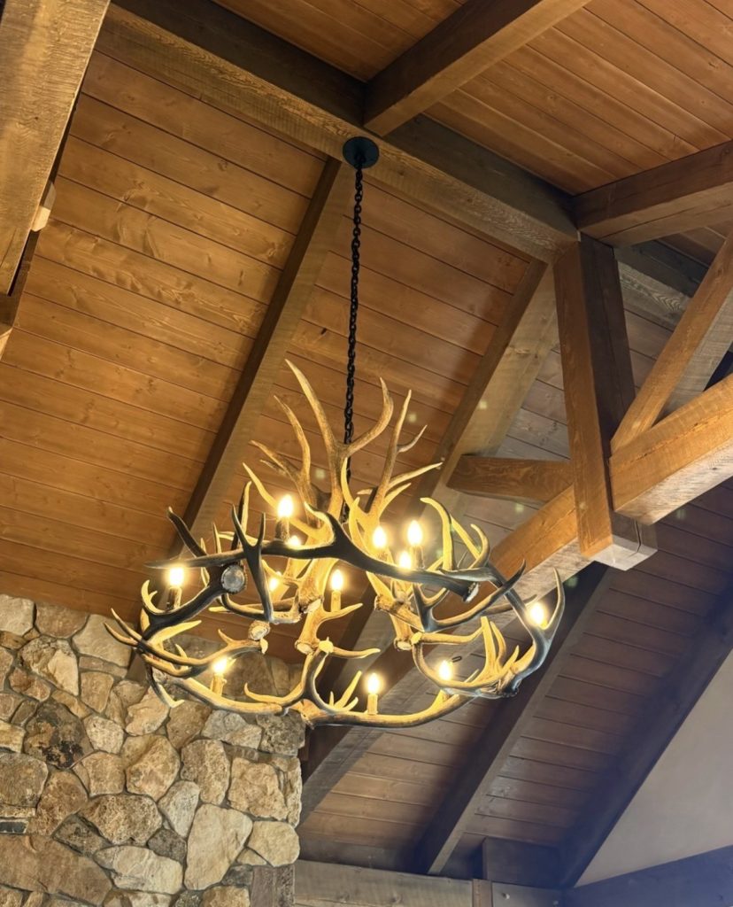 Big Sky commercial antler chandelier, lit, hanging from timber beams beside a stone fireplace