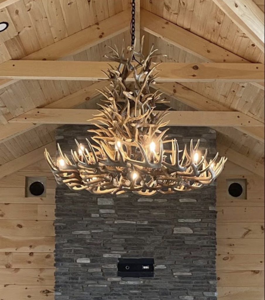 Lit six-tier whitetail shed antler chandelier hanging under timber beams in front of a stone chimney
