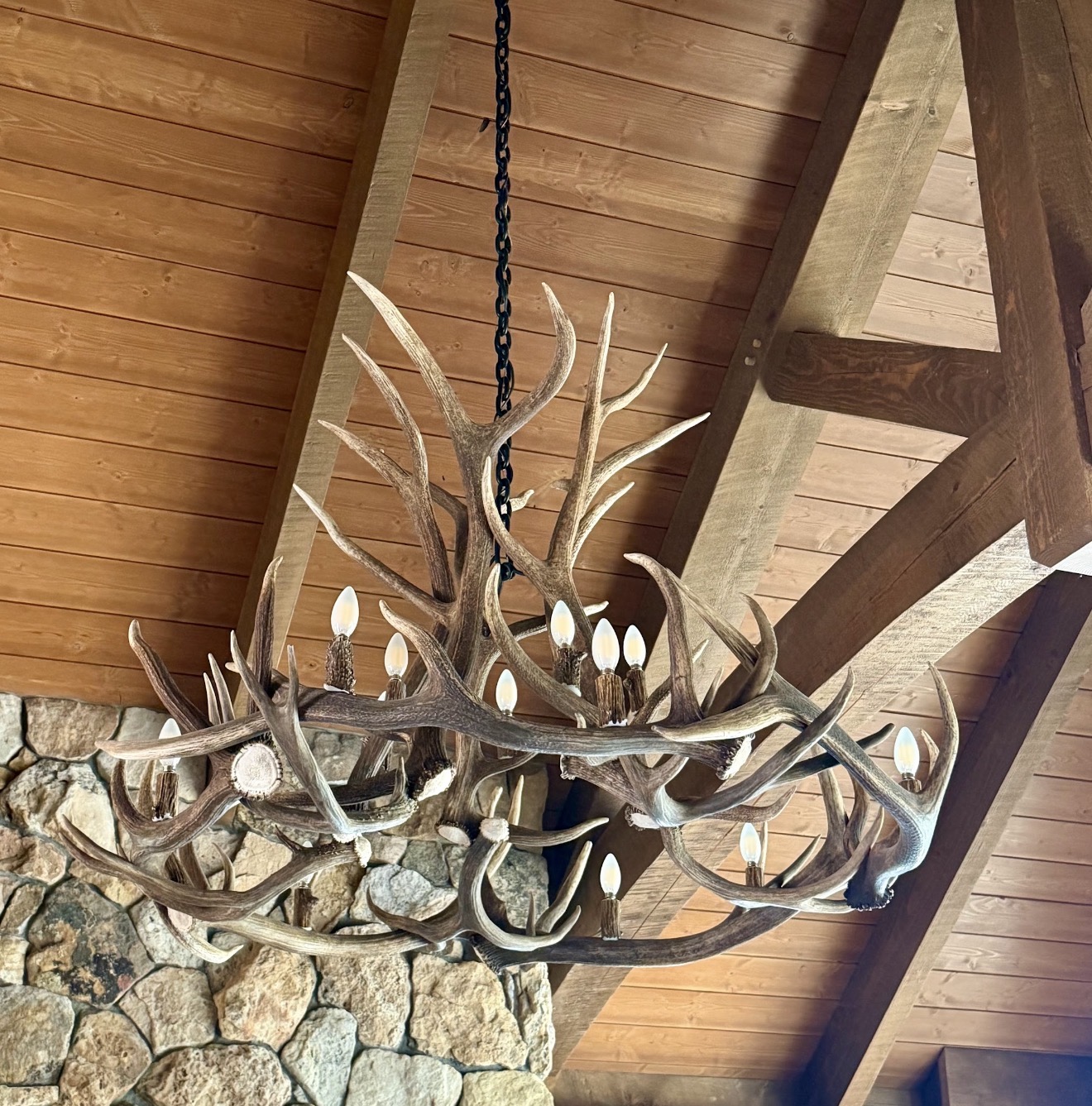 Antler chandelier hanging beneath a timber ceiling beside a stone wall.