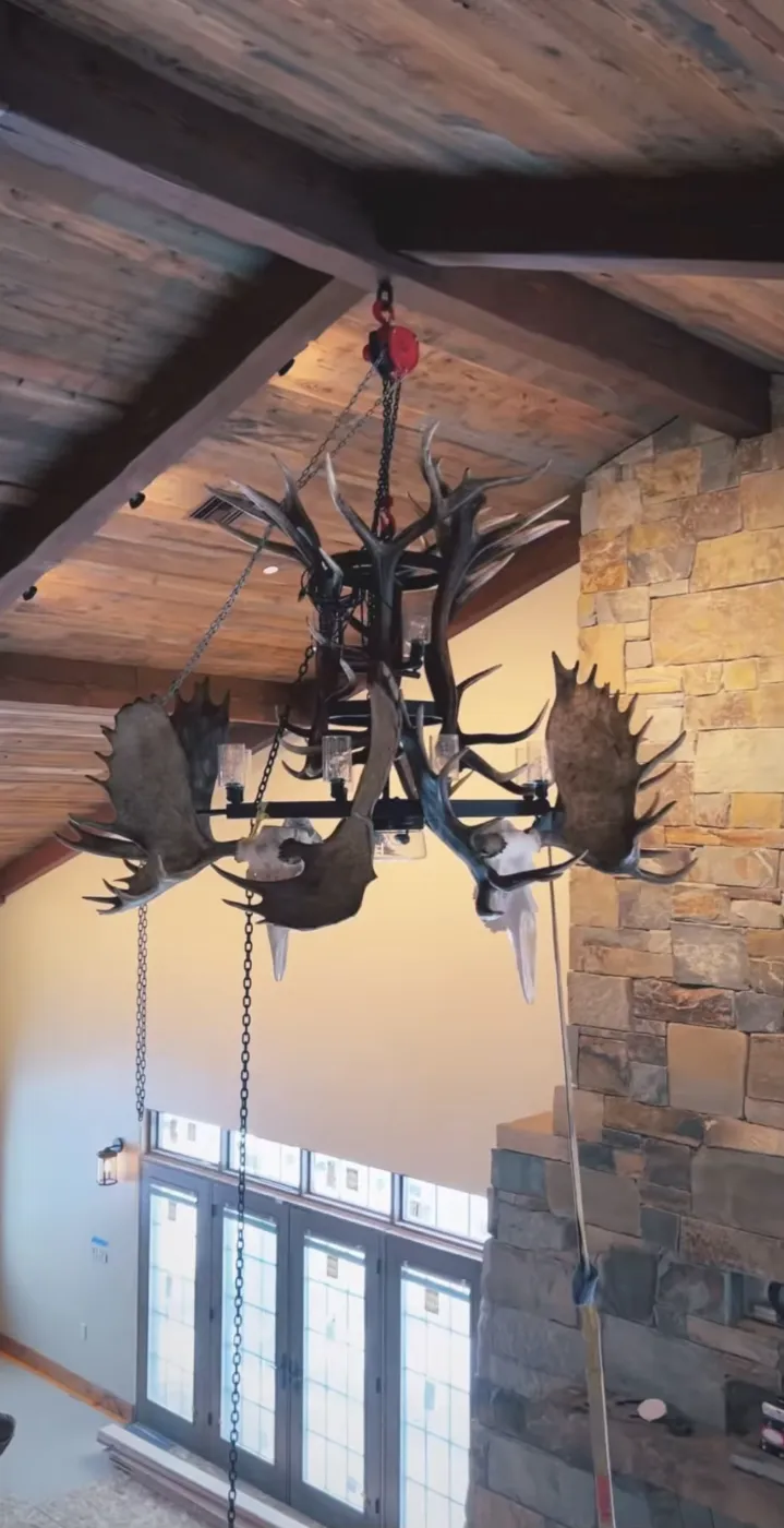 Antler chandelier with a dark metal frame mounted under a wood beam against a stone accent wall