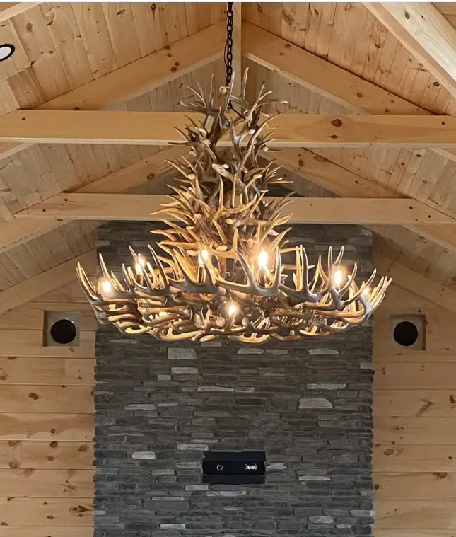 Large tiered round antler chandelier hanging in a vaulted wood-beamed room above a stone accent wall