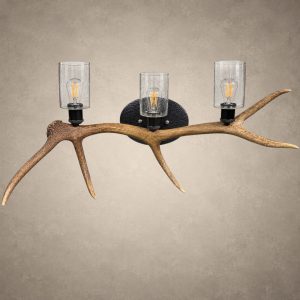 The Ridgeline elk antler 3-light wall light: a single shed elk antler mounted horizontally with three seeded glass cylinders and filament bulbs on a hammered steel back
