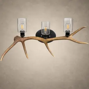 The Ridgeline elk antler 3-light wall light: a single shed elk antler mounted horizontally with three clear glass cylinders and filament bulbs on a hammered steel back